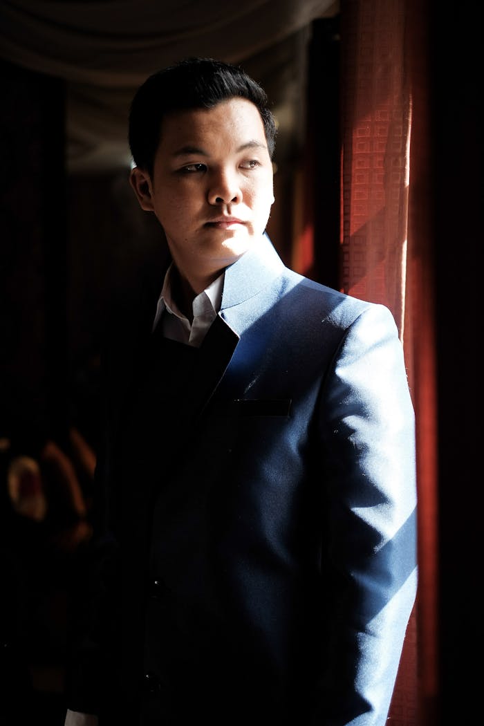 A thoughtful businessman in a stylish suit stands by a window, illuminated by sunlight.